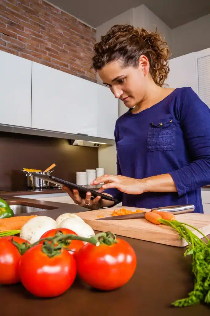 Elodie stands in a modern kitchen, looking at a tablet, surrounded by fresh vegetables on the counter.